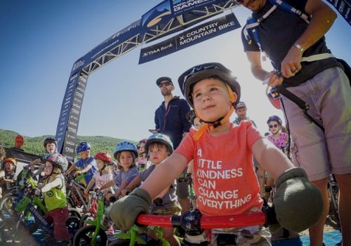 Children with helmets on their bikes at a starting line for a race, one in a red shirt that reads "A little kindness can change everything."