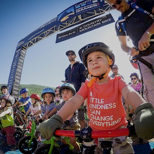 Children with helmets on their bikes at a starting line for a race, one in a red shirt that reads "A little kindness can change everything."