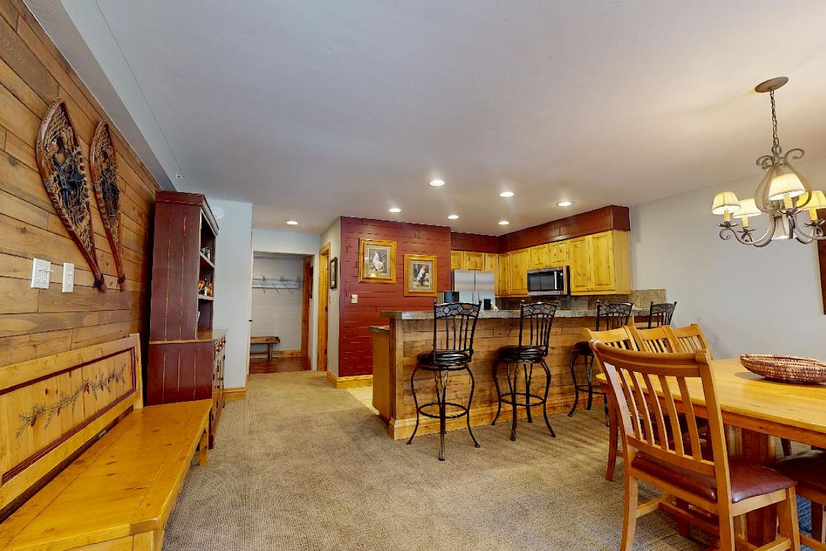 A cozy dining and kitchen area featuring wooden furniture, a chandelier, bar stools, and wall decorations. The space has a rustic, cabin-like feel.
