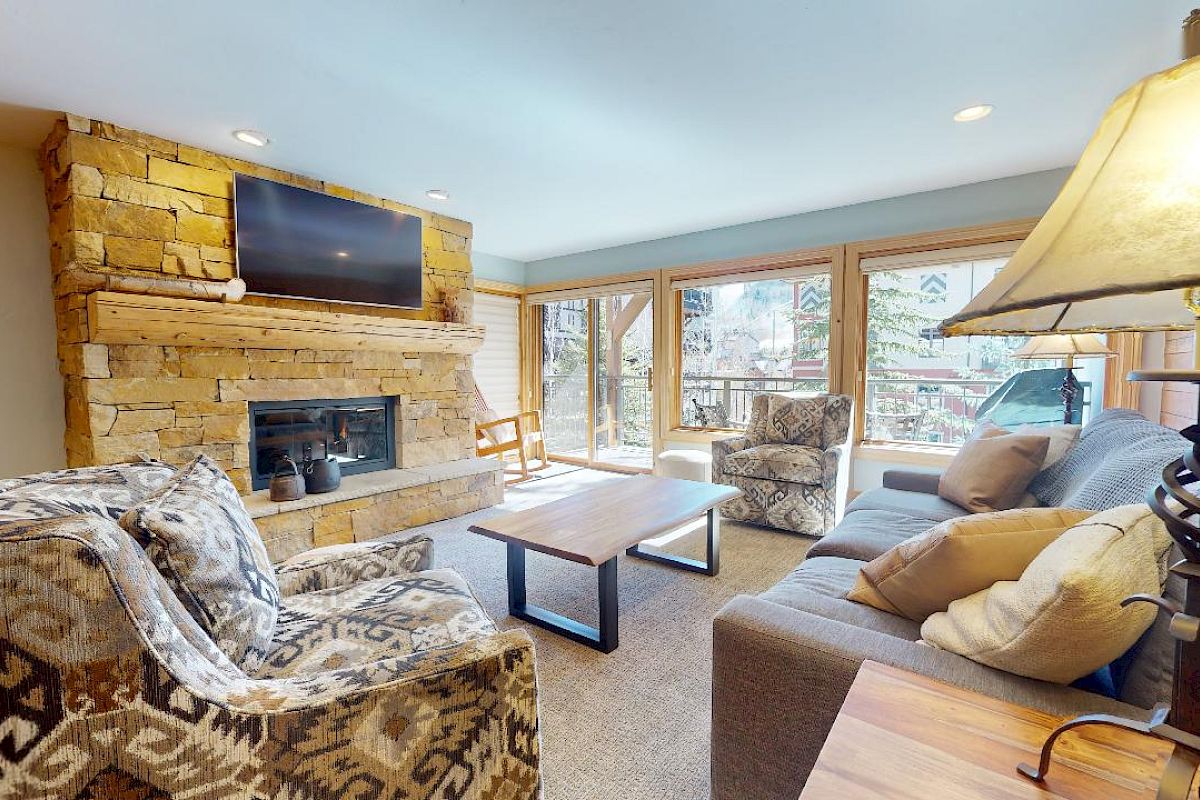 A cozy living room with a stone fireplace, wall-mounted TV, patterned armchairs, a couch with pillows, wooden coffee tables, and large windows.