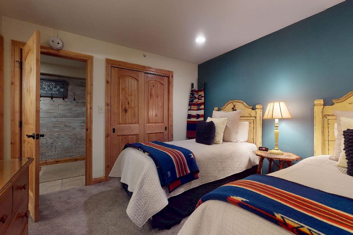 A cozy bedroom with two twin beds, a wooden bedside table with a lamp, a wooden dresser, and a closet with double doors.