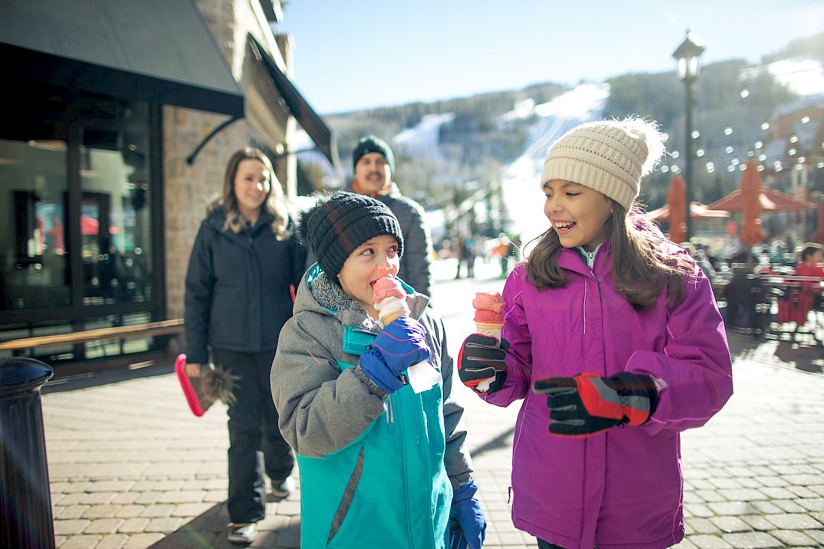 Two children in winter clothing enjoy ice cream while an adult couple follows behind them on a sunlit cobblestone street near a snowy mountain.