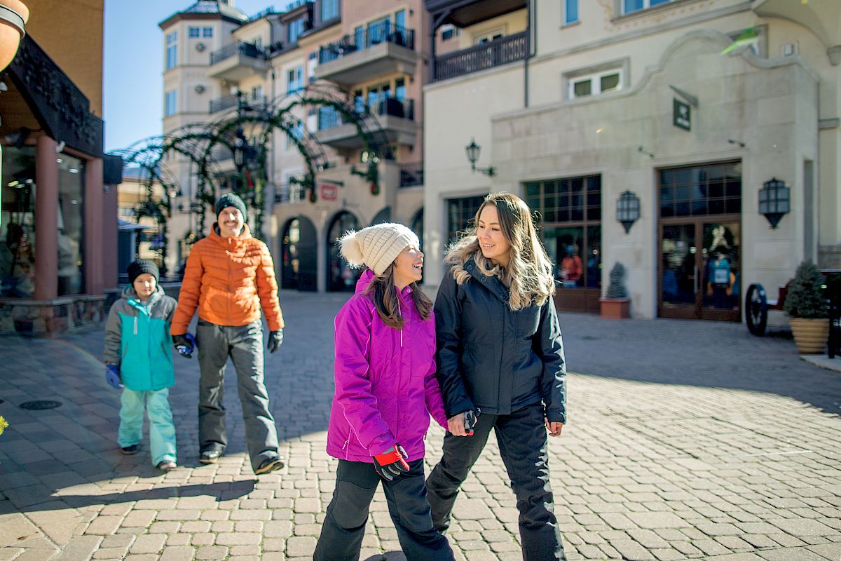 A group of people dressed in winter clothing walk through a scenic, cobblestone outdoor area with buildings in the background, enjoying the day.