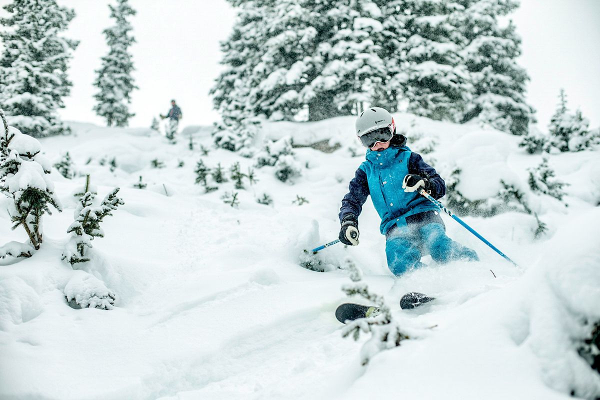 A person is skiing down a snowy slope surrounded by snow-covered trees, wearing blue ski gear, including a helmet and ski goggles, under a cloudy sky.