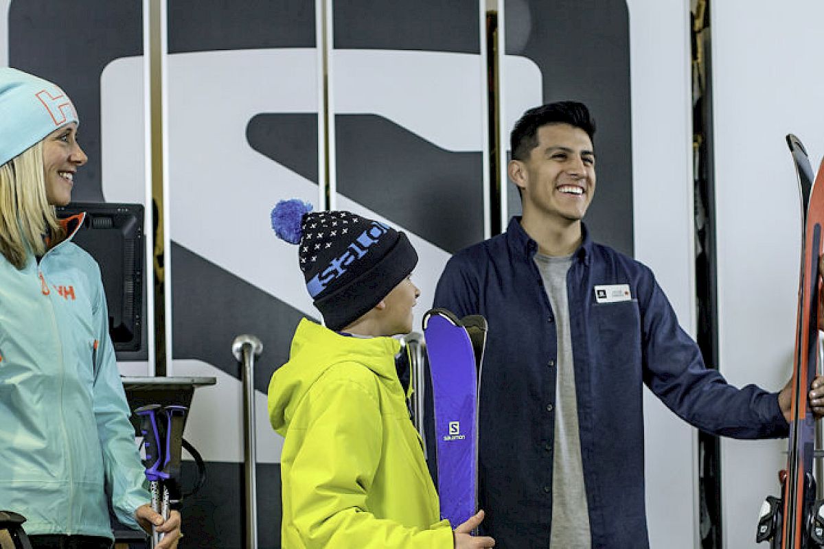 A group of people are in a ski rental shop, smiling and talking to an employee while holding skis and gear, preparing for a skiing activity.