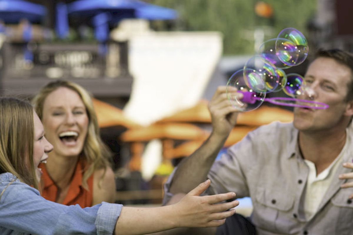 A group of four people are having fun outdoors, laughing as they play with soap bubbles. The background features orange umbrellas at an outdoor setting.