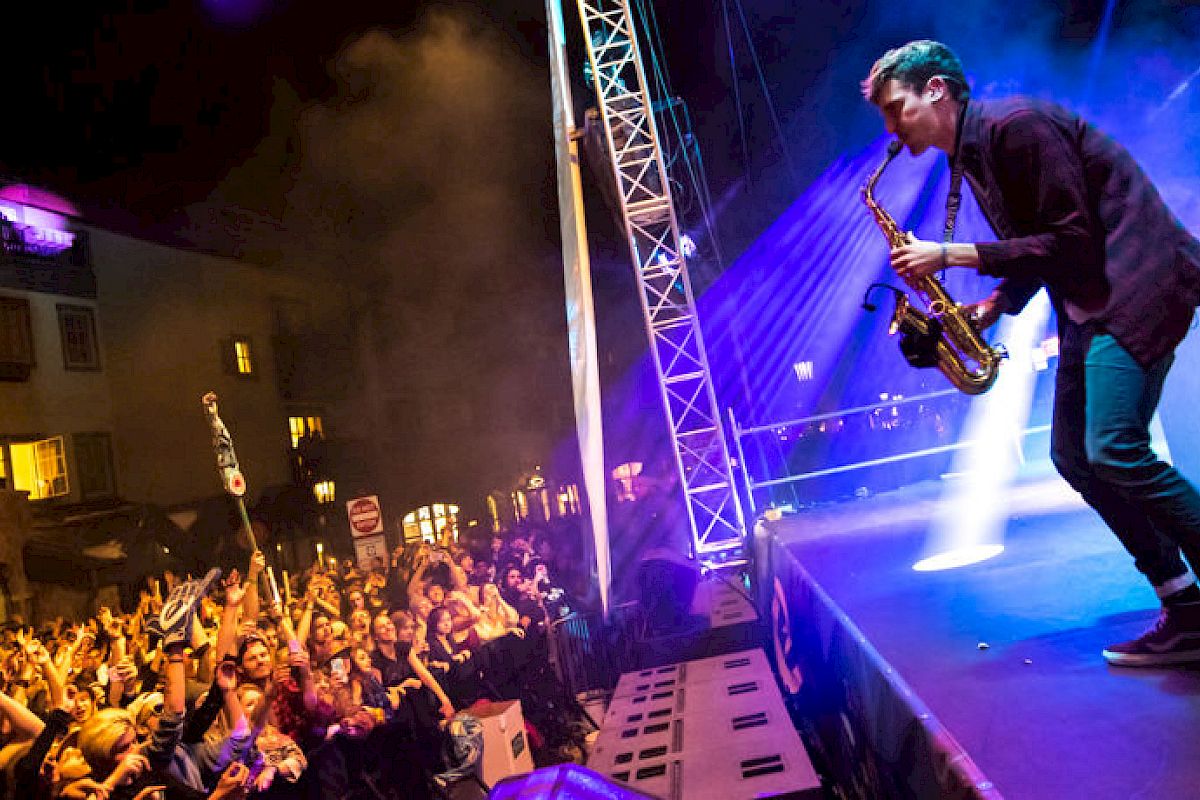 A musician is performing onstage with a saxophone, and a crowd of fans are cheering in front of a lit-up outdoor venue during a night concert.