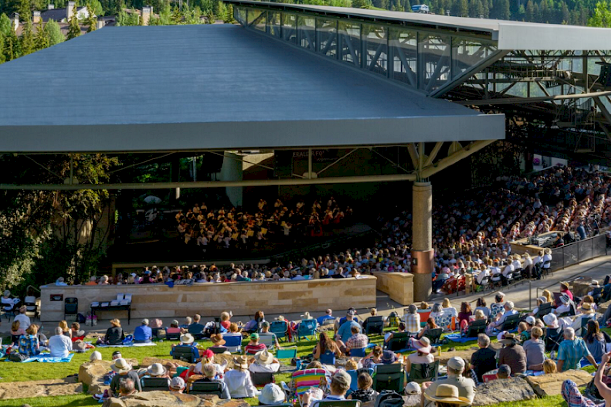 A large outdoor concert venue with a seated audience, surrounded by lush greenery and a forested backdrop, under a canopy structure.