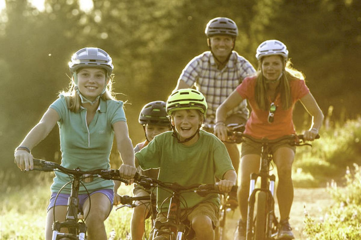 A group of people, including two adults and three children, are riding bicycles through a sunlit, grassy area, all wearing helmets.