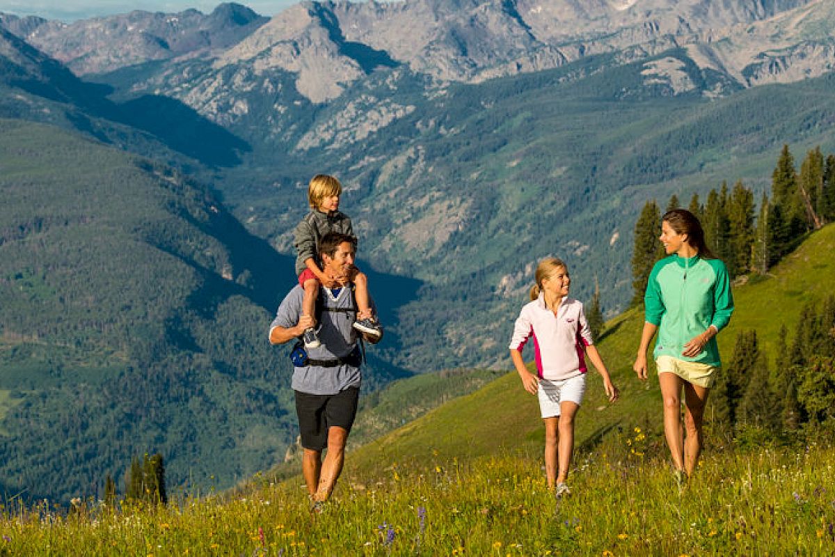 A family of four hikes through a lush green mountain meadow, with the stunning backdrop of mountains. One child is carried on an adult's shoulders.