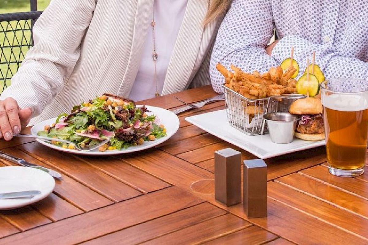 Two people seated at an outdoor wooden table with a salad, fried dish, wine, and beer. No faces shown.