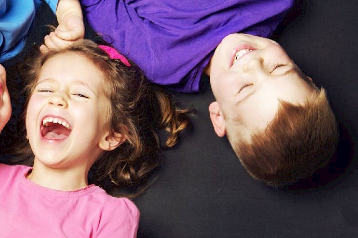 Three children are lying on the ground, heads together, laughing joyfully, and wearing colorful clothes.