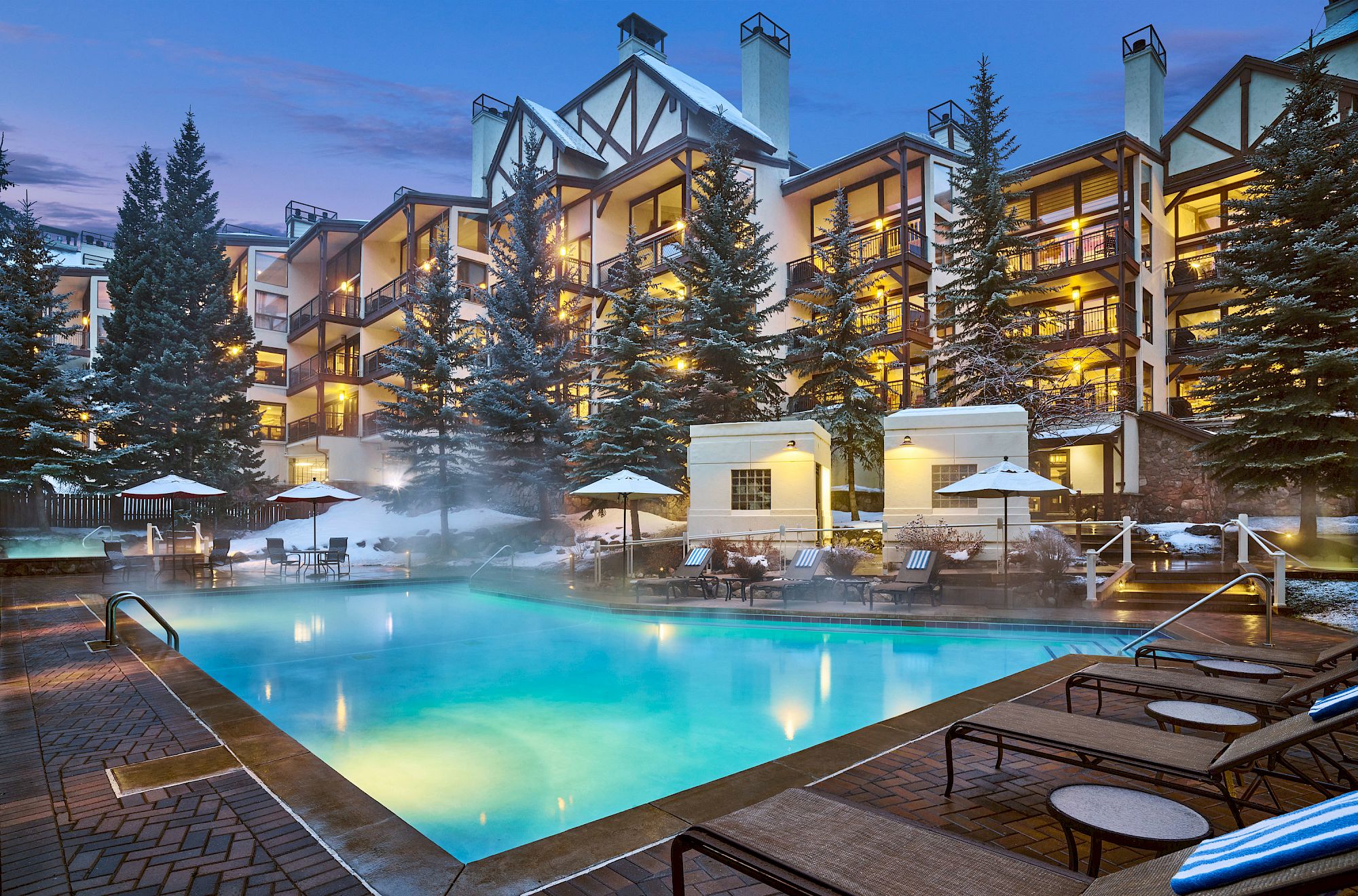 The image shows a lit outdoor pool surrounded by lounge chairs and trees, with a multi-story building in the background against a twilight sky.