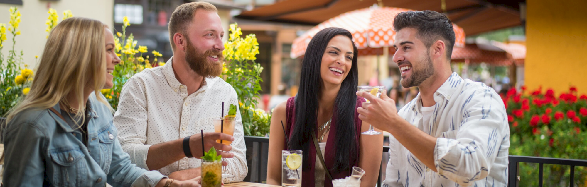 Four people are seated outdoors, enjoying drinks and laughing, with flowers and a patio in the background.