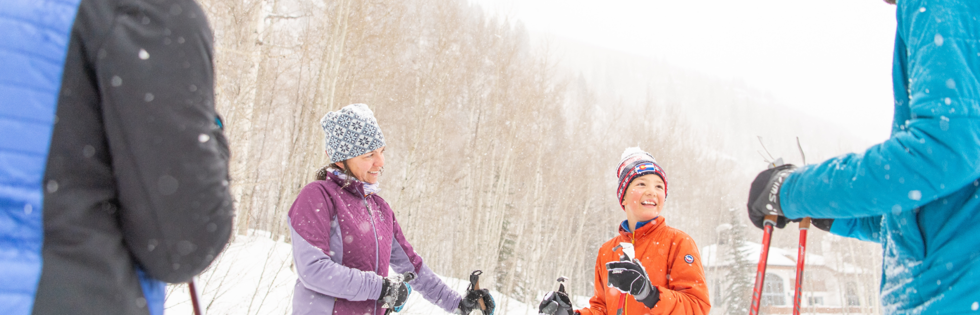 Group of people in winter clothing, holding ski poles, smiling and talking in a snowy outdoor setting with trees in the background.