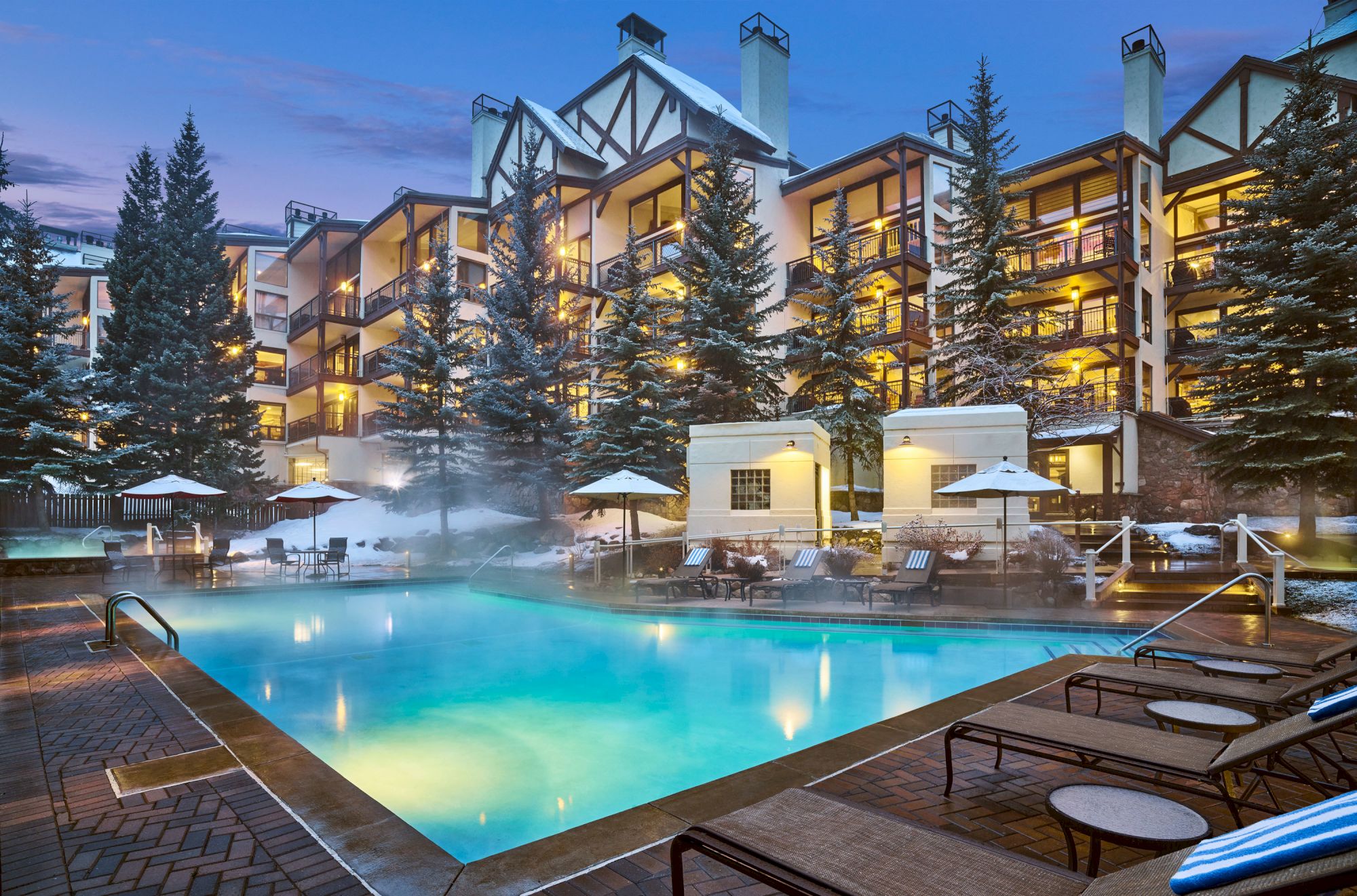 The image shows an outdoor heated pool surrounded by lounge chairs and umbrellas, with a multi-story building and tall pine trees in the background.
