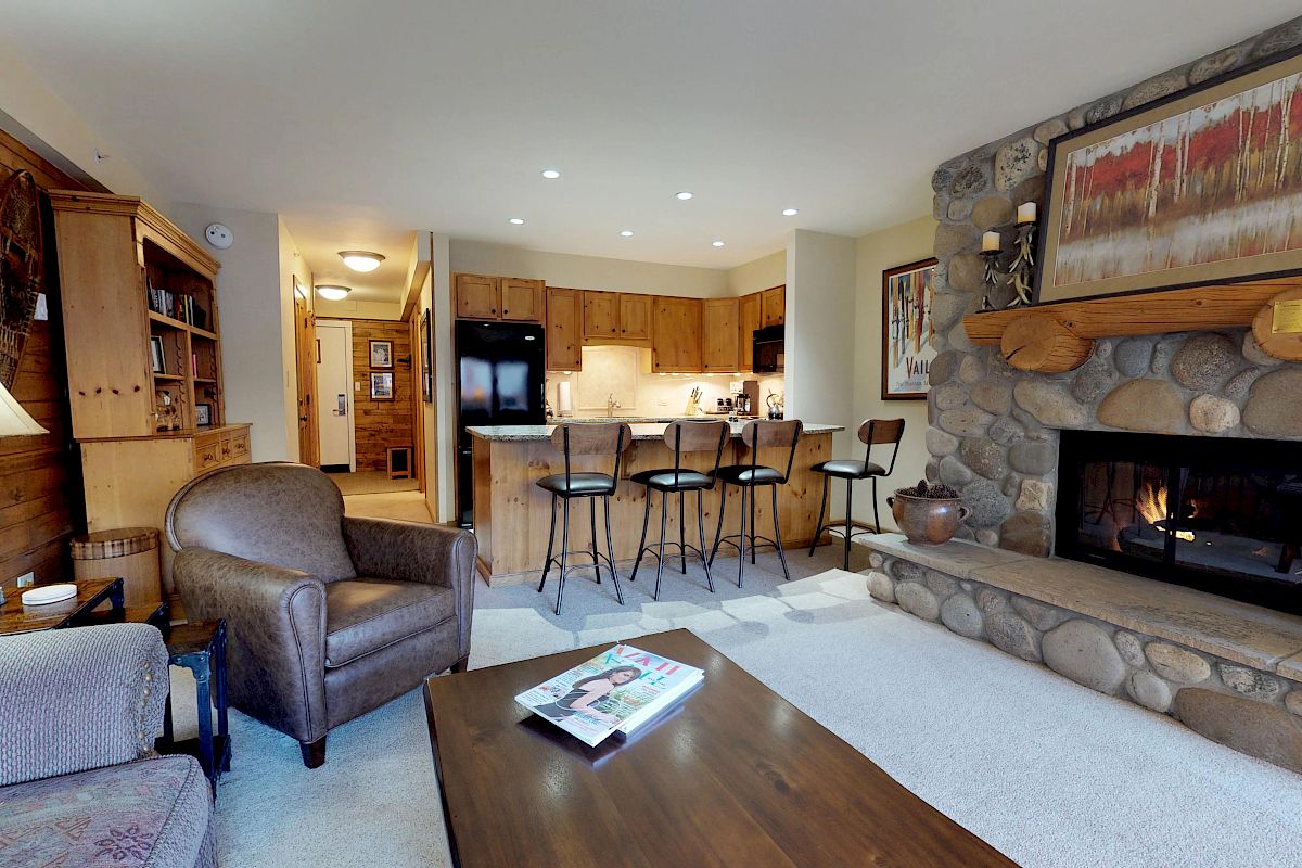 A cozy living room with a stone fireplace, wooden furniture, a kitchen with a breakfast bar, and comfortable seating.