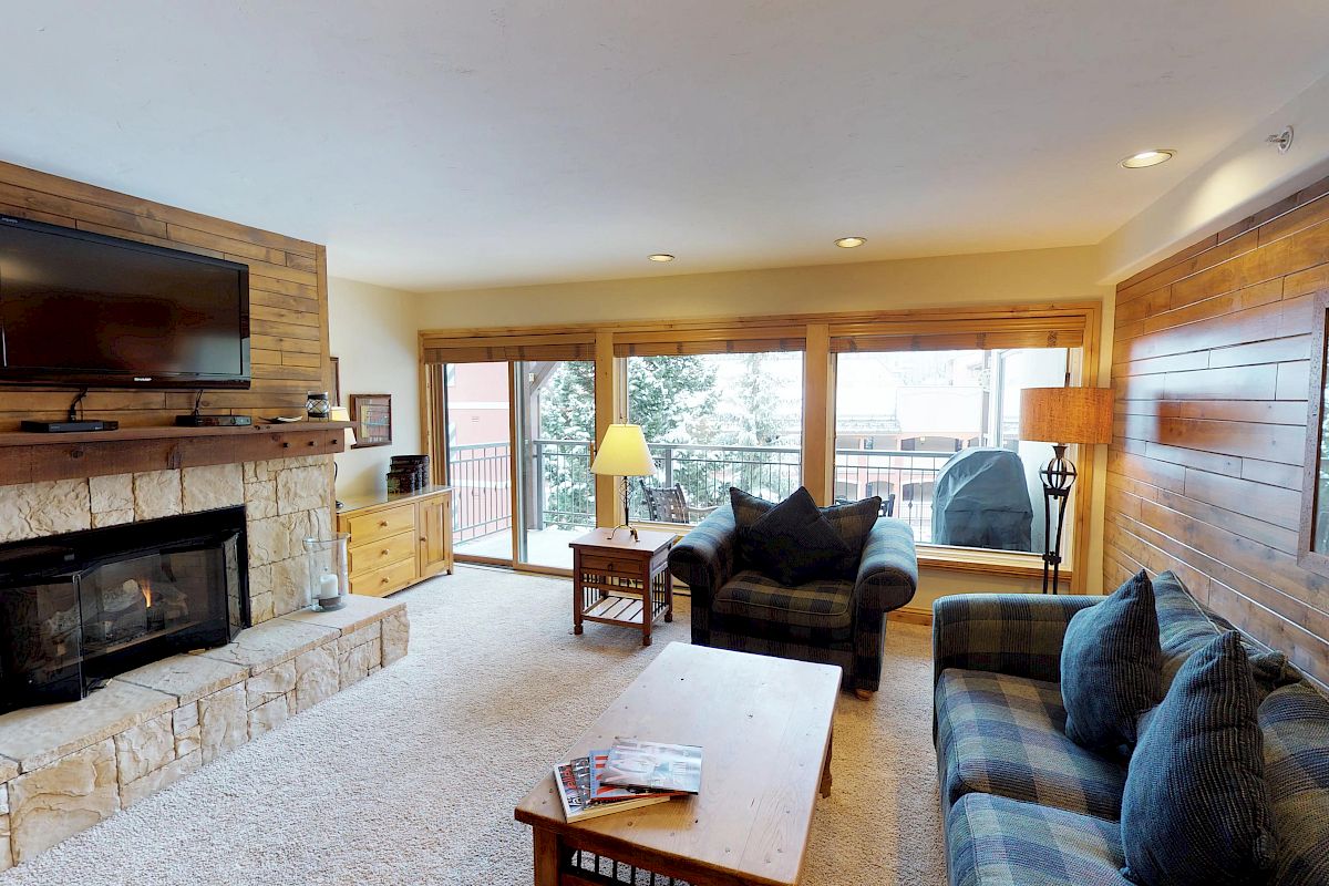 A cozy living room features a stone fireplace, mounted TV, plaid couch, armchair, wood coffee table, and large windows overlooking a snowy landscape.