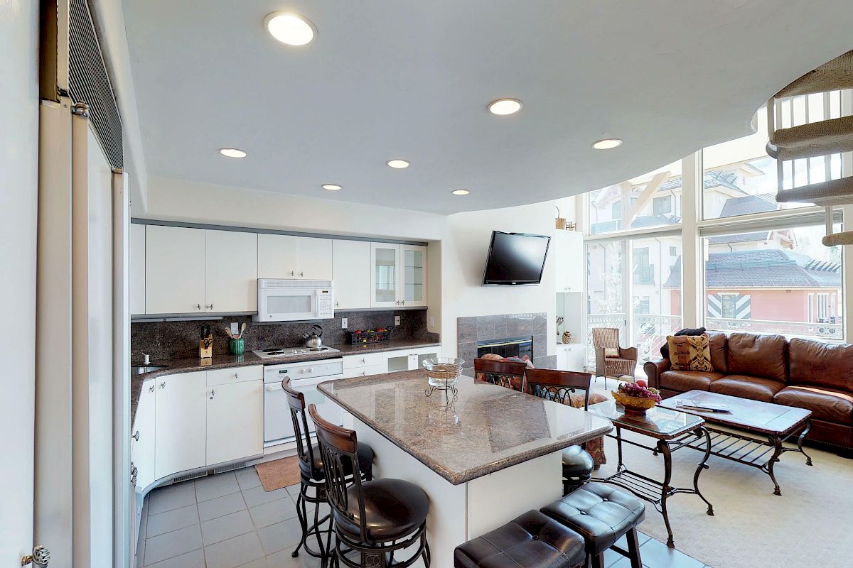 A modern kitchen and living area with a granite island, bar stools, sofa, coffee table, wall-mounted TV, fireplace, and spiral staircase.