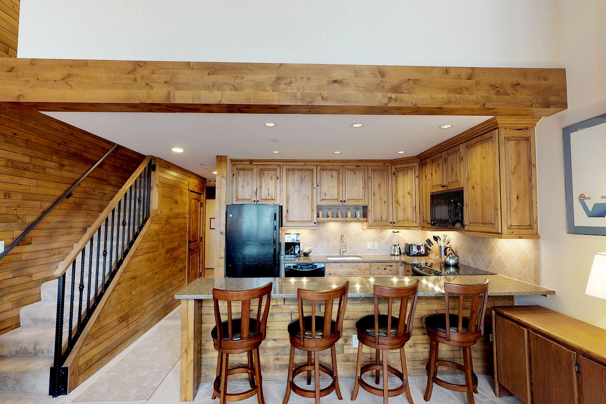 The image depicts a rustic kitchen with wooden cabinets, a bar counter with four stools, a black refrigerator, and a staircase leading upwards.