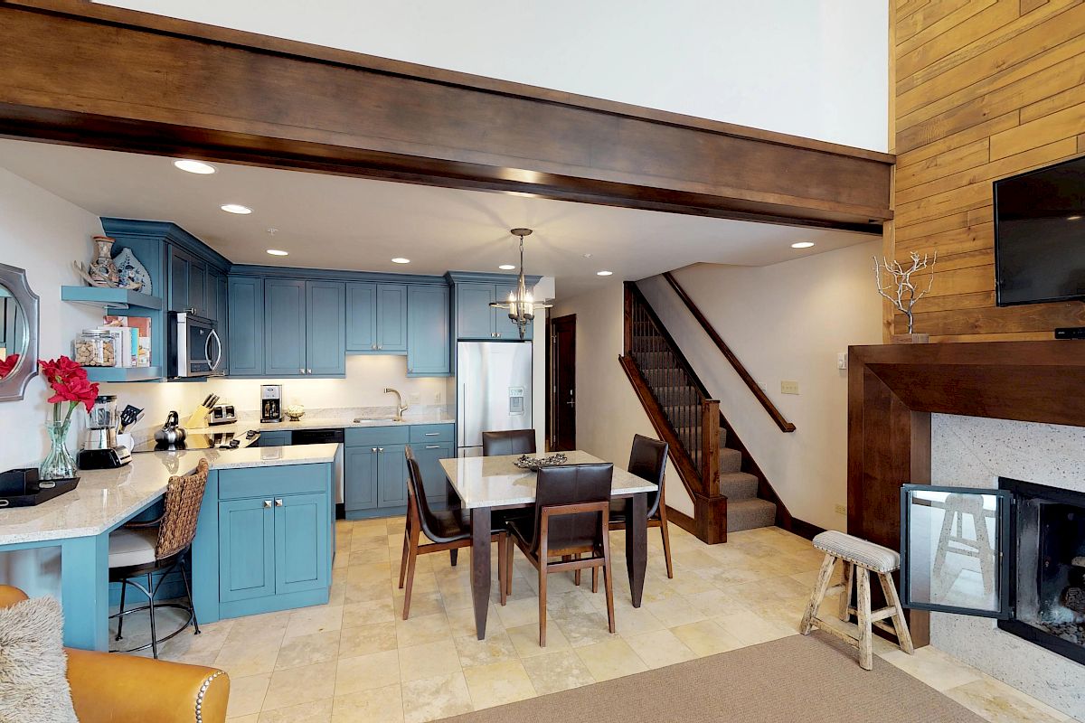 This image shows a modern kitchen and dining area with blue cabinets, a wooden ceiling, a cozy fireplace, and a staircase leading upstairs.