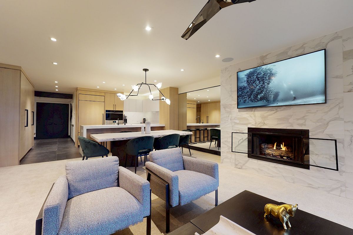 Contemporary living room with a fireplace, wall-mounted TV, modern furniture, a kitchen island with bar stools, and a dining area in the background.