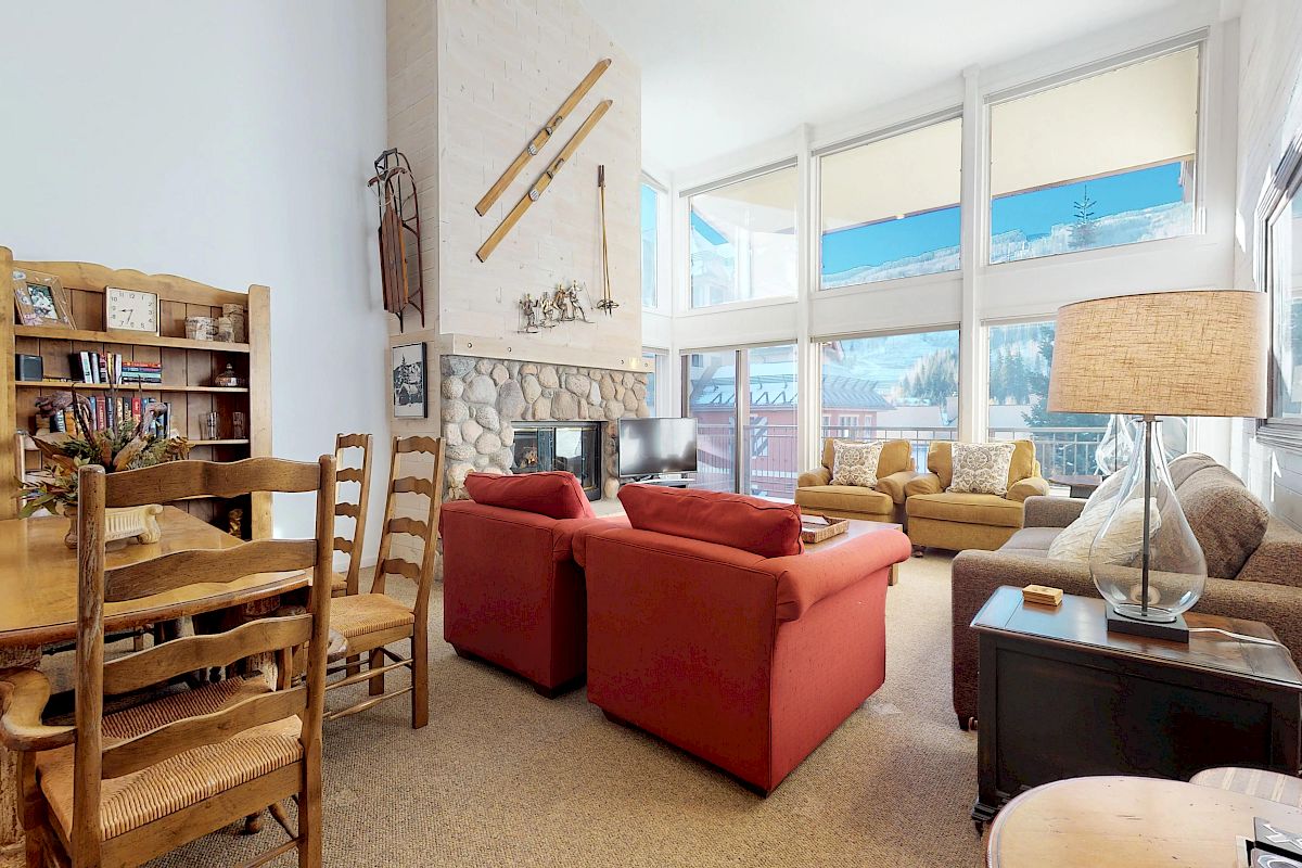 A cozy living room with red armchairs, a fireplace, large windows, and wooden furniture. Skis decorate the wall above the fireplace.