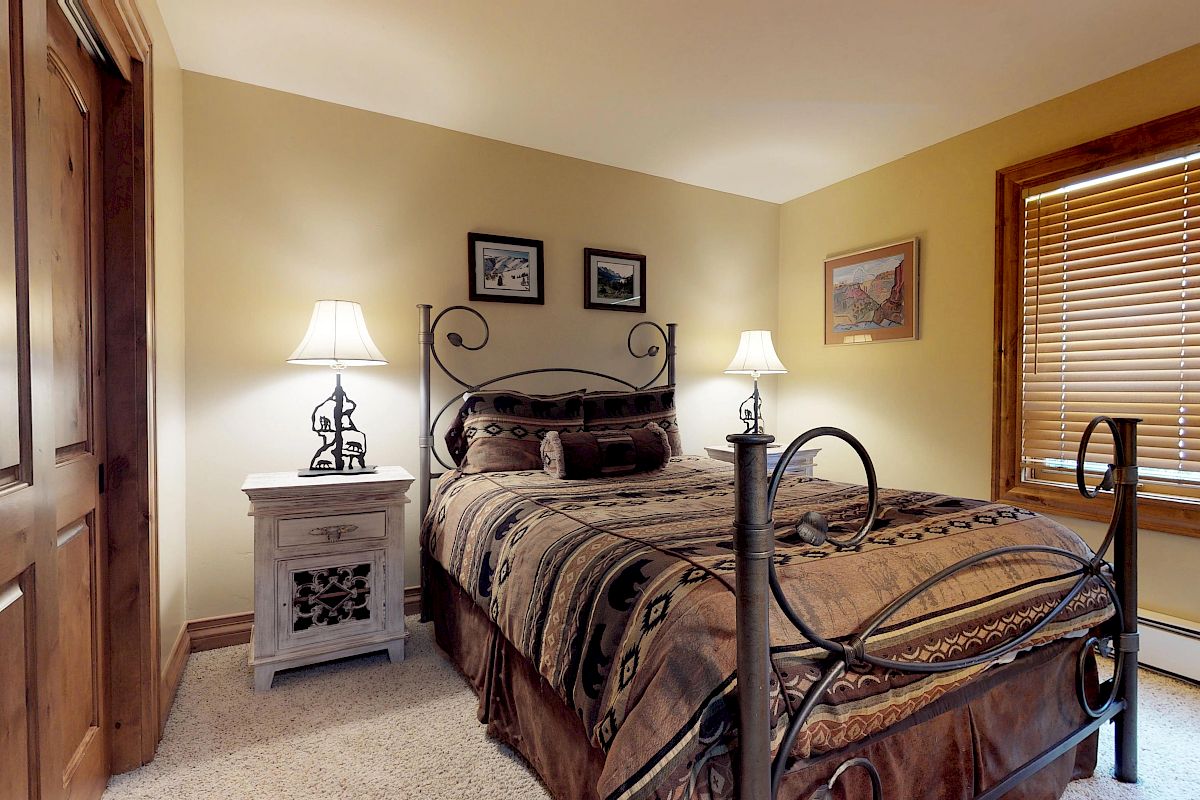 A cozy bedroom features a wrought iron bed frame, patterned bedding, two nightstands with lamps, wall art, a wooden door, and a window with blinds.