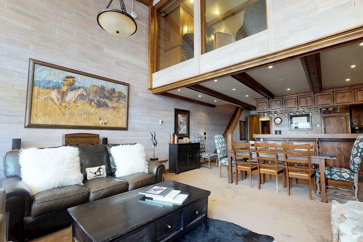 A spacious living room with high ceilings, a leather sofa, fluffy pillows, a painting, a dining table, and a kitchen area with wooden cabinets.