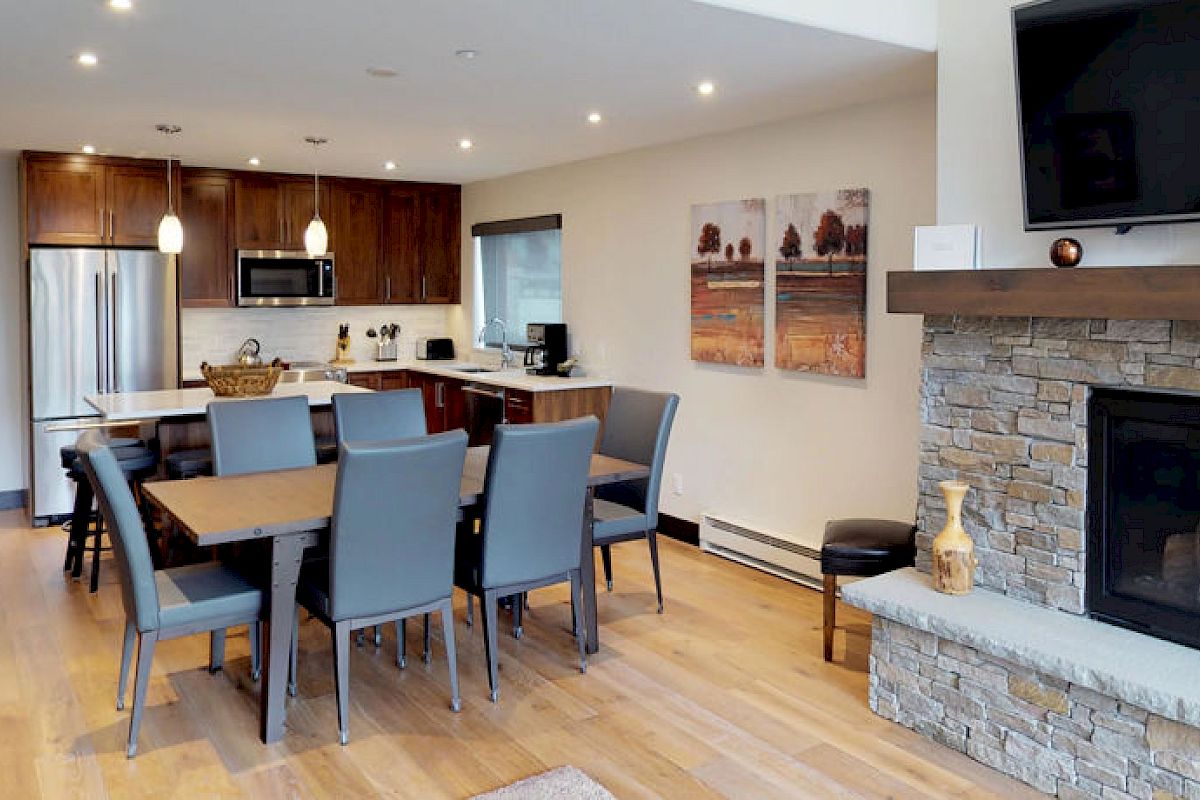 Modern living area with wooden floors, open kitchen, dining table, stone fireplace, and a TV. Contemporary staircase on the left side of the room.