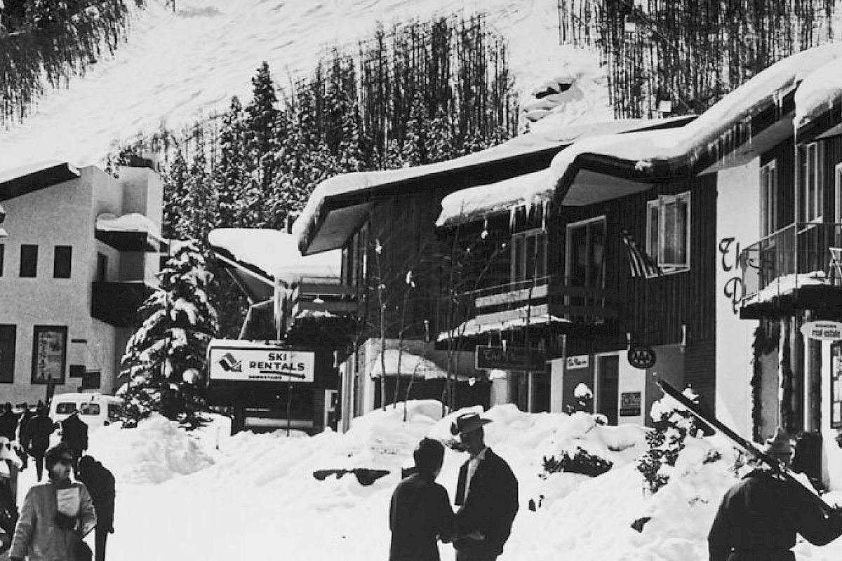 A snowy street scene in an alpine village with snow-covered buildings; people are walking, including some carrying ski gear.