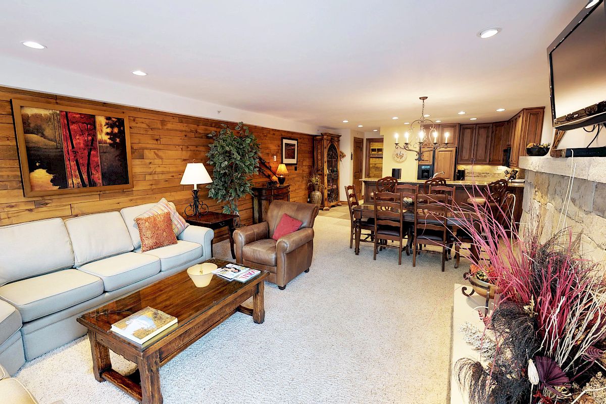 A cozy living room with a sectional sofa, armchair, wooden coffee table, wall art, plants, dining area, and an open kitchen in the background.