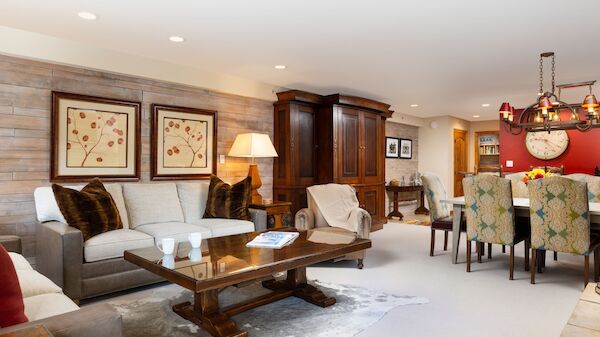 The image shows a cozy living area with a sofa, coffee table, artwork, a dining table, and warm wood accents throughout.