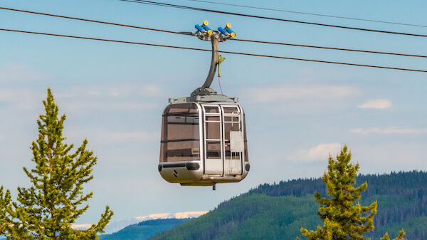 The image shows a cable car suspended on wires, moving above a forested area with mountains in the background.