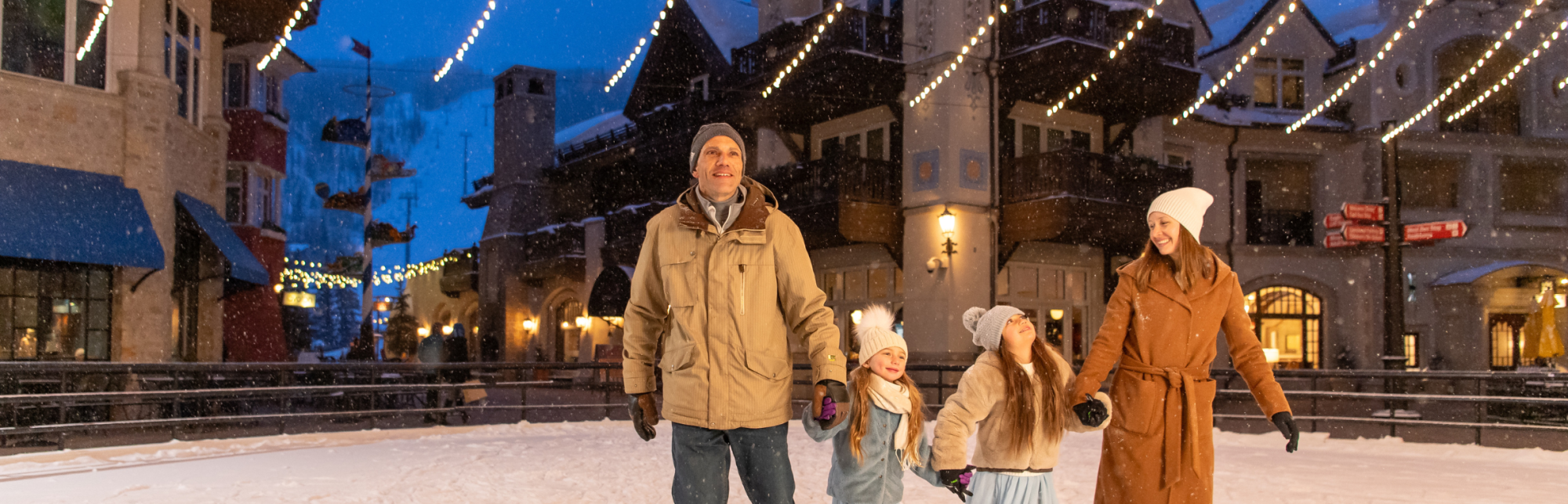 A family of four in winter clothing stands on a snowy street with festive lights and charming buildings in the background.