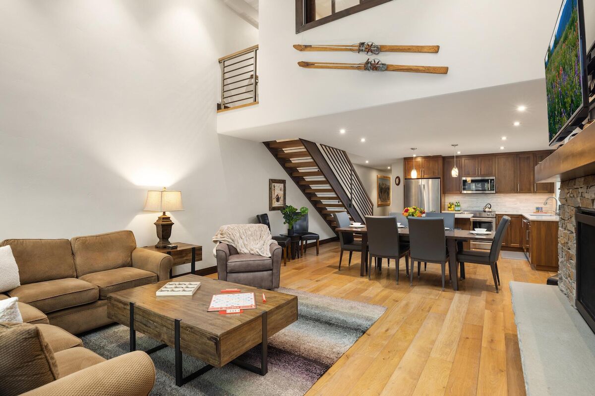 A bright open-concept living room with tan couches, a wooden coffee table, and a modern kitchen/dining area in the background. It features a staircase and decorative skis on the wall, plus warm lighting.