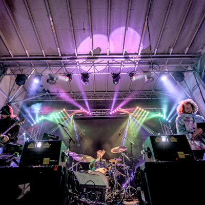 A band is performing on stage with colorful lights and smoke effects, showcasing a drummer, guitarist, and keyboardist in action, surrounded by equipment.