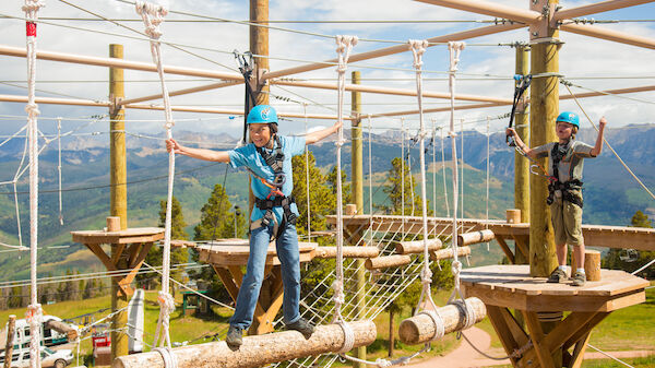 Two children wearing helmets and harnesses navigate a ropes course outdoors, with a scenic mountain backdrop. They appear to be enjoying the activity.