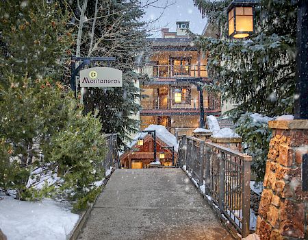 A snow-covered pathway with railings leads to buildings with warm lights, surrounded by trees. A sign labeled "Montaneros" is visible on the left.