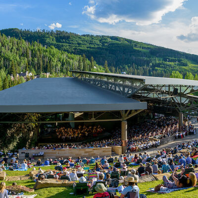 An outdoor amphitheater set in a lush, mountainous area with a large audience seated on the grass, enjoying a live performance.