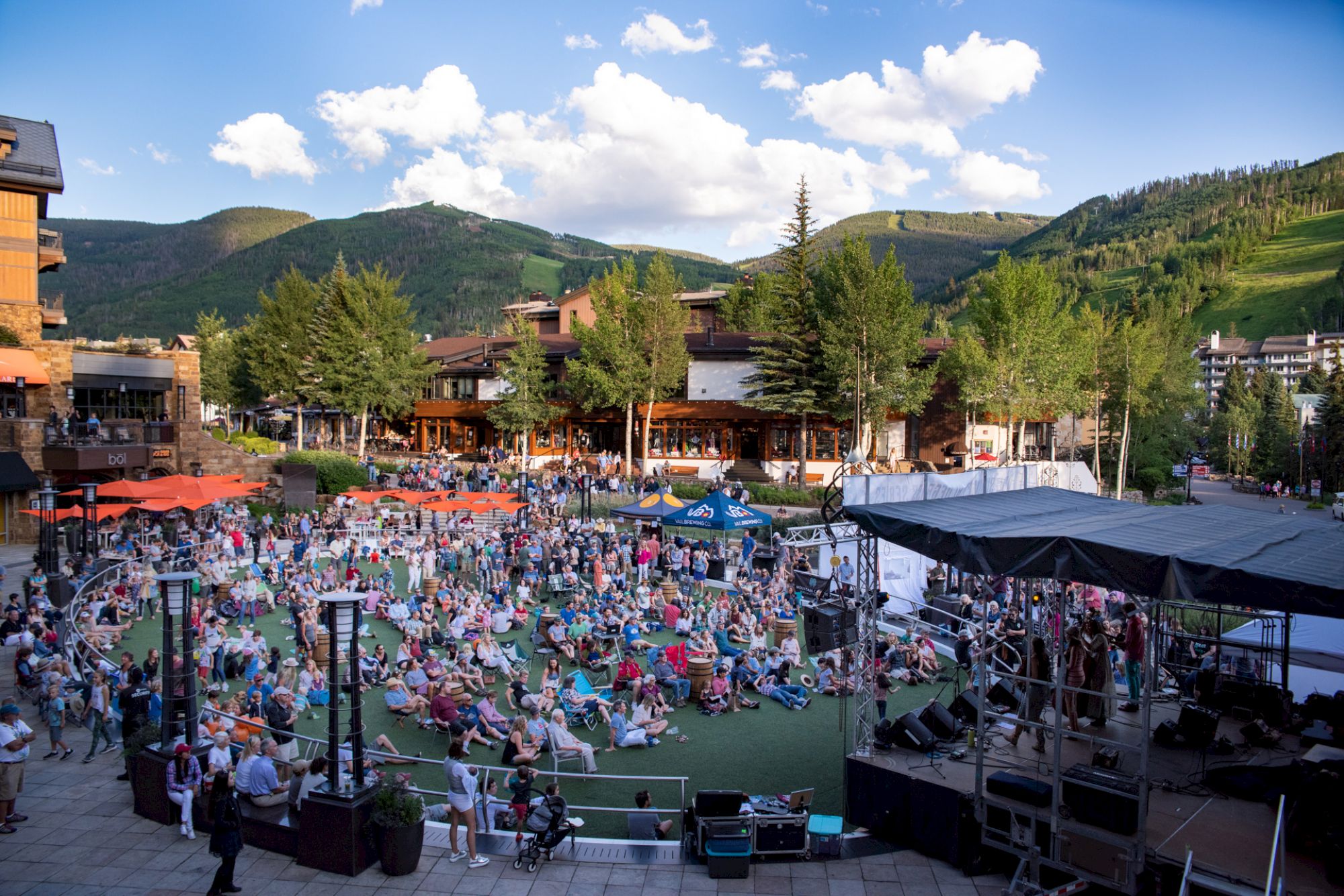 People are gathered outdoors on a lawn with mountains and trees in the background, enjoying a live performance on a stage.