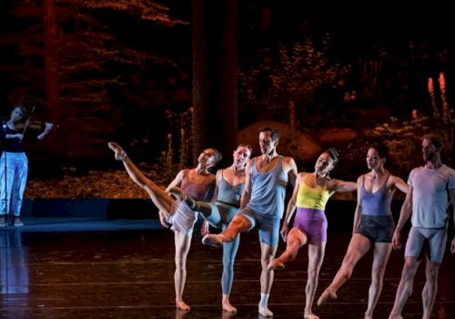 Dancers are performing on stage, with a violinist playing in the background, set against a scenic backdrop resembling a forest.