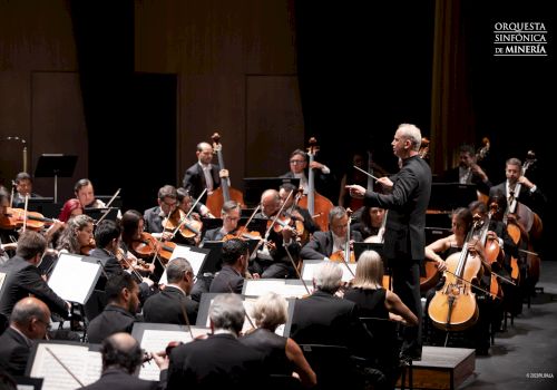 A conductor leads an orchestra during a performance; musicians play various instruments, with "Orquesta Sinfónica de Minería" logo in the upper right.