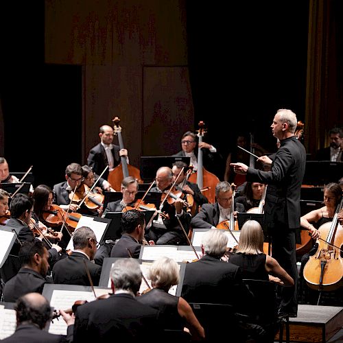 A conductor leads an orchestra during a performance; musicians play various instruments, with "Orquesta Sinfónica de Minería" logo in the upper right.