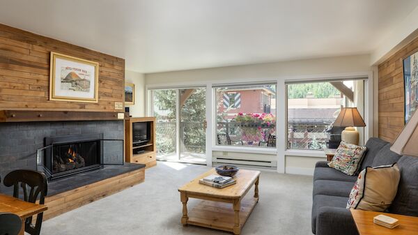 Cozy living room with a stone fireplace, wooden accents, large windows, a gray sofa, coffee table, and a few chairs, bright and inviting.
