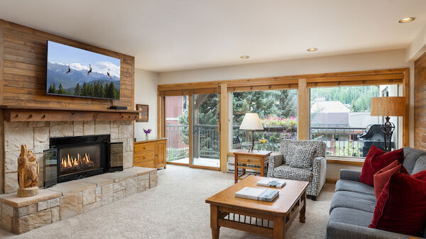 The image shows a cozy living room with a fireplace, comfortable furniture, and large windows offering a view of nature.