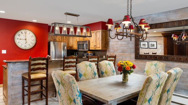 A rustic modern kitchen-dining area with a wooden table, upholstered chairs, red accent wall, bar seating, chandelier, and flower centerpiece.