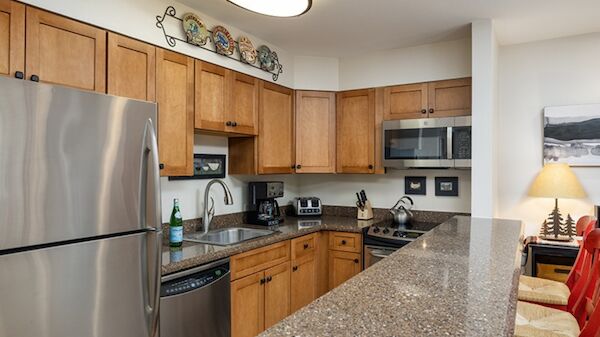 A modern L-shaped kitchen with wooden cabinets, stainless steel fridge, microwave, dishwasher, granite countertop bar, and red chairs in a dining area.