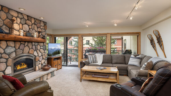 A cozy living room with a stone fireplace, plush sofas, a TV, and a balcony view. Vintage skis are decoratively mounted on the wall.