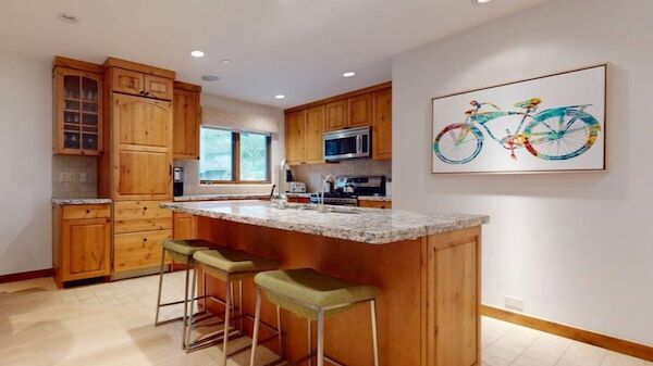 The image shows a modern kitchen with wooden cabinets, a granite countertop, and bar stools, featuring a colorful bicycle artwork.